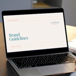 May include: A silver laptop shows a document titled "Brand Guidelines" with the text "That Branding Coach" and the logo "The Branding Coach". The screen displays the version and date. The laptop is on a wooden surface.