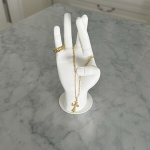 K&ouml;nnte beinhalten: Wei&szlig;e Keramik-Handskulptur mit einer goldenen Kettenkette und einem goldenen Ring. Die Kette hat einen Kreuzanh&auml;nger.