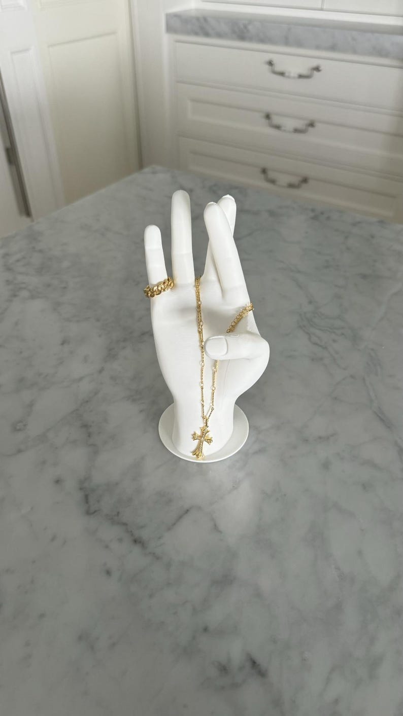 K&ouml;nnte beinhalten: Wei&szlig;e Keramik-Handskulptur mit einer goldenen Kettenkette und einem goldenen Ring. Die Kette hat einen Kreuzanh&auml;nger.