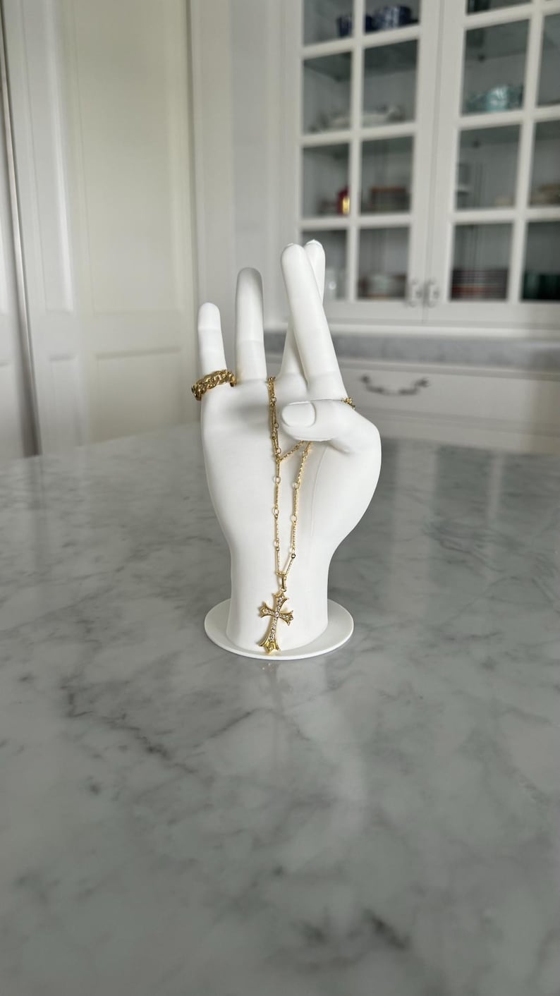 K&ouml;nnte beinhalten: Wei&szlig;e Keramik-Handskulptur mit einer goldenen Kettenkette und einem Kreuzanh&auml;nger. Die Hand h&auml;lt die Kette und das Kreuz h&auml;ngt herunter. Die Hand steht auf einem wei&szlig;en Sockel.