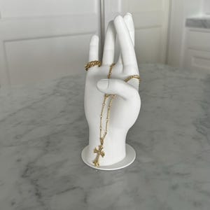 K&ouml;nnte beinhalten: Wei&szlig;e Keramik-Handskulptur mit einer goldenen Kettenkette und einem Kreuzanh&auml;nger. Die Hand h&auml;lt die Kette und das Kreuz h&auml;ngt herunter.