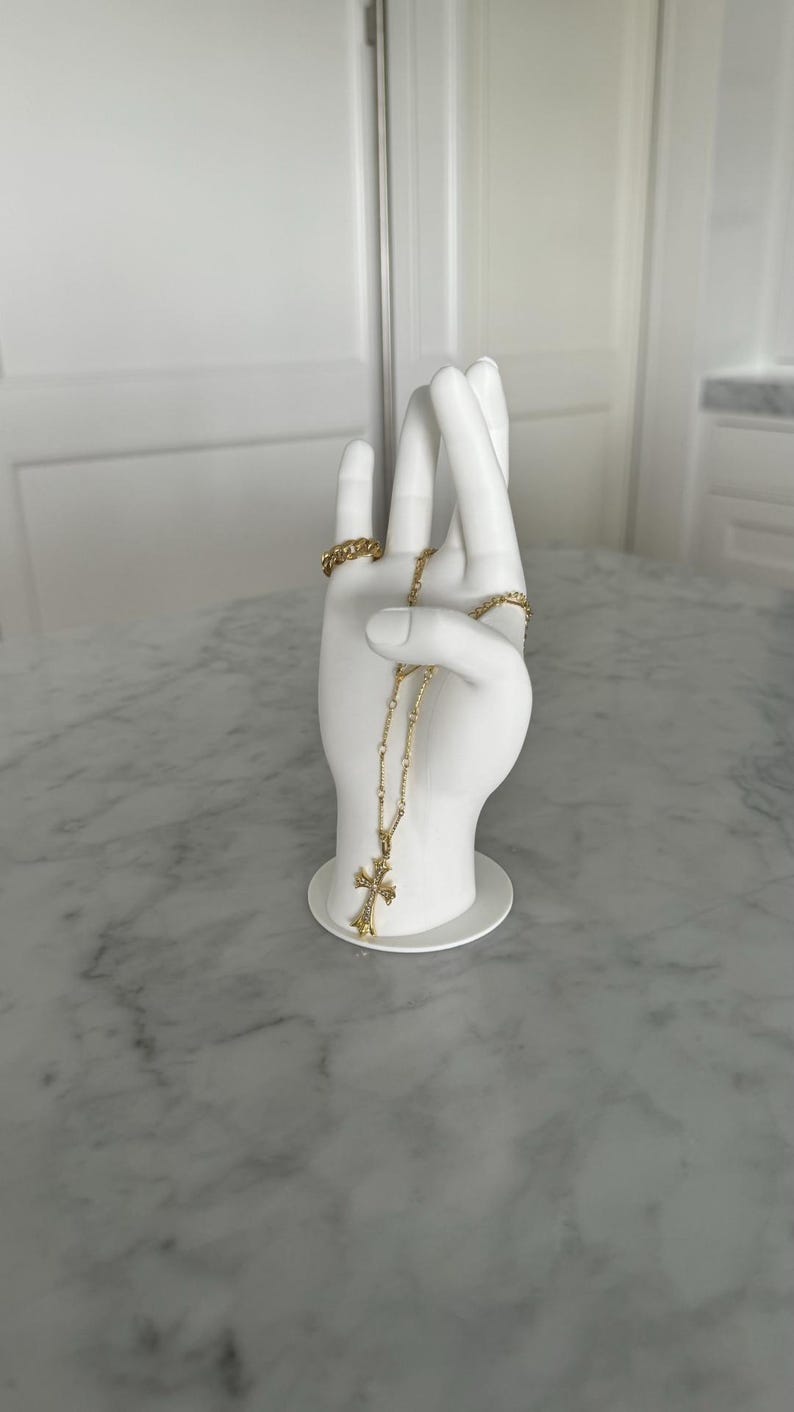 K&ouml;nnte beinhalten: Wei&szlig;e Keramik-Handskulptur mit einer goldenen Kettenkette und einem Kreuzanh&auml;nger. Die Hand h&auml;lt die Kette und das Kreuz h&auml;ngt herunter.