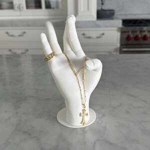 K&ouml;nnte beinhalten: Wei&szlig;er Keramik-Hand-Ring- und Halskettenhalter mit einer goldenen Kettenkette und einem goldenen Kreuzanh&auml;nger.