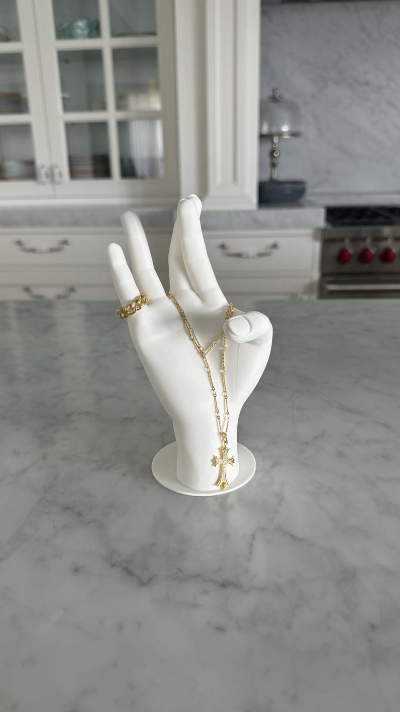 K&ouml;nnte beinhalten: Wei&szlig;er Keramik-Hand-Ring- und Halskettenhalter mit einer goldenen Kettenkette und einem goldenen Kreuzanh&auml;nger.