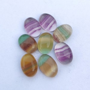 May include: Eight oval fluorite gemstones in various colours, including purple, green, yellow, and brown, with banded patterns. The polished stones are arranged on a white surface. Each stone is approximately 2-3 cm in length.