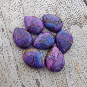 May include: Seven teardrop-shaped purple, blue, and gold gemstone cabochons. The cabochons have a marbled pattern and are arranged in a circle on a wooden surface.