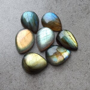 Puede incluir: Una colección de siete piedras preciosas de labradorita en forma de lágrima en varios tonos de verde, azul y dorado. Las piedras son lisas y pulidas, con un brillo nacarado.