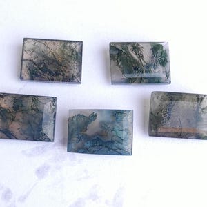 Puede incluir: Cinco cabujones rectangulares de ágata musgosa con una variedad de patrones verdes, grises y marrones. Las piedras son translúcidas y tienen un acabado pulido.