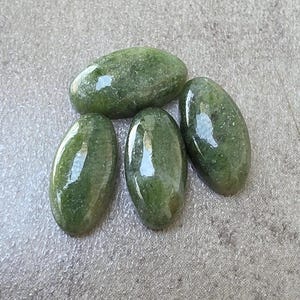 Könnte beinhalten: Vier ovale, polierte grüne Edelsteine. Die Steine haben eine glatte, glänzende Oberfläche und variieren leicht in der Größe. Die Edelsteine ​​liegen auf einer strukturierten, grauen Oberfläche.