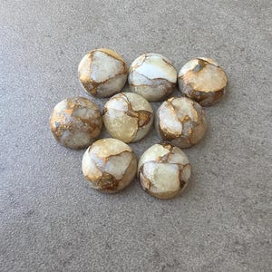 May include: Eight round, polished stones with a cream-colored base and intricate gold veining. The stones are arranged on a gray surface, showcasing their unique patterns and textures. These stones could be used for jewelry making or decorative purposes.