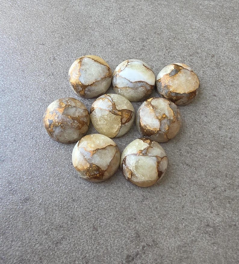 May include: Eight round, polished stones with a cream-colored base and intricate gold veining. The stones are arranged on a gray surface, showcasing their unique patterns and textures. These stones could be used for jewelry making or decorative purposes.