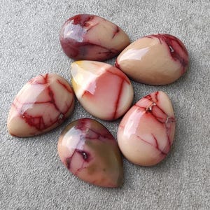 以下が含まれることがあります： 6つの磨かれた涙型の宝石。石は、鮮やかな赤い静脈が全体に走る、クリーミーなベージュのベースカラーをしています。磨かれた表面は光を反射し、複雑な模様を際立たせています。宝石は灰色の表面に配置されています。