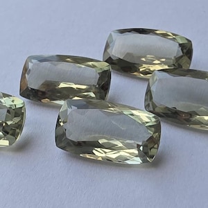 Puede incluir: Cinco gemas rectangulares facetadas con un tono verde oliva pálido. Las gemas están dispuestas sobre una superficie blanca, cada una mostrando múltiples facetas que capturan la luz. Las piedras tienen una apariencia pulida y translúcida.