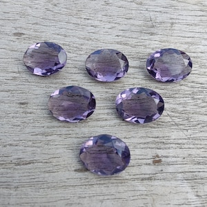 May include: Six oval-shaped amethyst gemstones, each with a deep purple hue, are arranged on a light grey wooden surface.