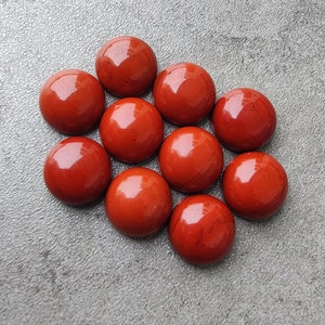 以下が含まれることがあります： 滑らかで磨かれた表面を持つ赤いジャスパーの石が10個集められています。石は灰色の背景に円形に配置されています。