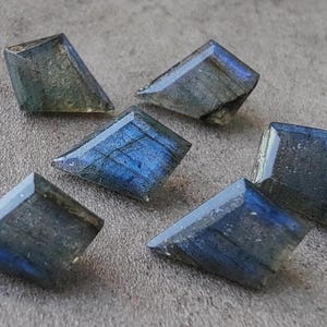 May include: Six faceted labradorite gemstones with blue and green iridescence. The gemstones are cut into a trapezoid shape and are displayed on a textured grey surface. The stones have a polished, reflective surface.