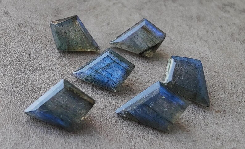 May include: Six faceted labradorite gemstones with blue and green iridescence. The gemstones are cut into a trapezoid shape and are displayed on a textured grey surface. The stones have a polished, reflective surface.