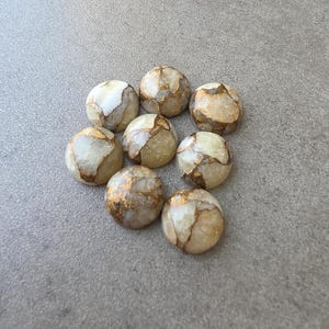 May include: Eight round, polished stones with a cream and gold color scheme. The stones have a marbled appearance with gold veining throughout. The stones are arranged in a circular pattern on a gray surface.