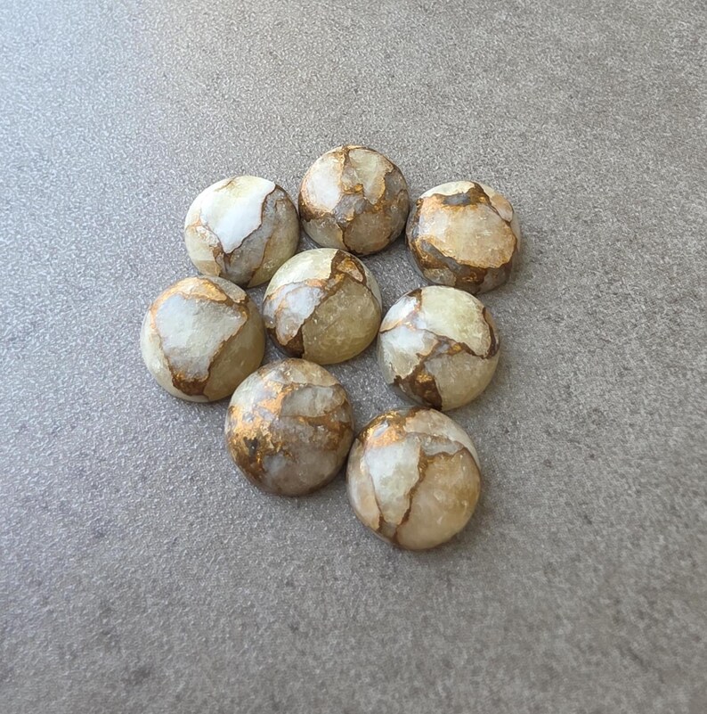 May include: Eight round, polished stones with a cream and gold color scheme. The stones have a marbled appearance with gold veining throughout. The stones are arranged in a circular pattern on a gray surface.