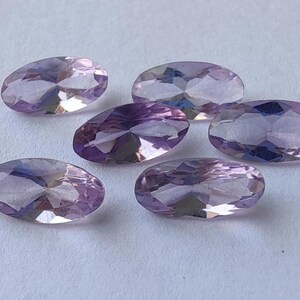 Peut inclure: Six pierres précieuses ovales et facettées, dans des tons lavande et violets, sont disposées sur une surface blanche. Les pierres ont une surface polie et réfléchissante, captant la lumière et affichant des facettes internes.