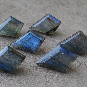 May include: Six faceted labradorite gemstones in shades of blue, grey, and green. The gemstones are cut into a rectangular shape with angled edges, displaying a subtle shimmer.