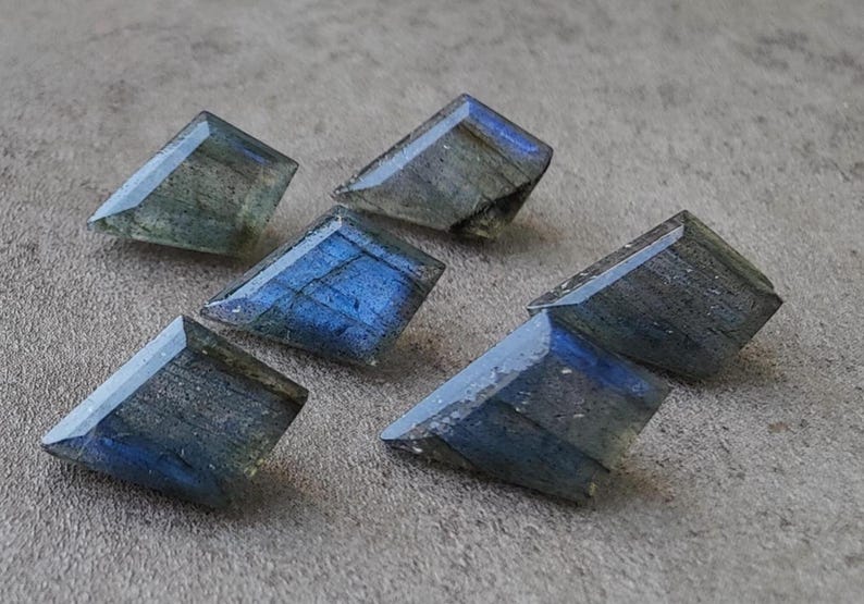 May include: Six faceted labradorite gemstones in shades of blue, grey, and green. The gemstones are cut into a rectangular shape with angled edges, displaying a subtle shimmer.