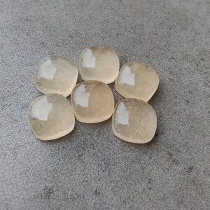 May include: Six translucent, rounded, square-shaped stones with a pale yellow hue are arranged on a gray surface. Each stone has a glossy, reflective surface, catching the light. The stones appear smooth and polished, suggesting they may be gemstones or decorative elements.