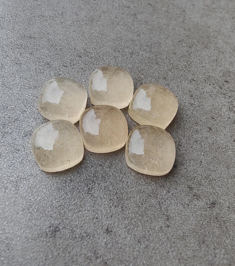 May include: Six translucent, rounded, square-shaped stones with a pale yellow hue are arranged on a gray surface. Each stone has a glossy, reflective surface, catching the light. The stones appear smooth and polished, suggesting they may be gemstones or decorative elements.