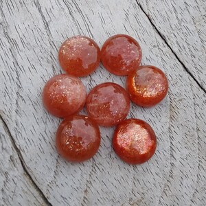 May include: Seven round, orange-red sunstone cabochons with a sparkly, iridescent finish. The stones are arranged in a circle on a wooden surface.