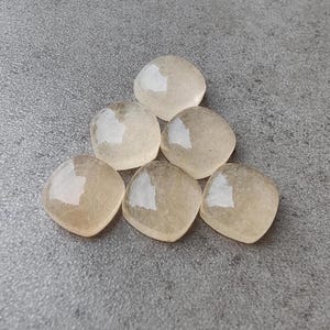 May include: Six light beige, square-shaped stones are arranged on a gray surface. Each stone has a rounded shape with a slightly reflective, white patch on one side. The stones appear smooth and translucent.