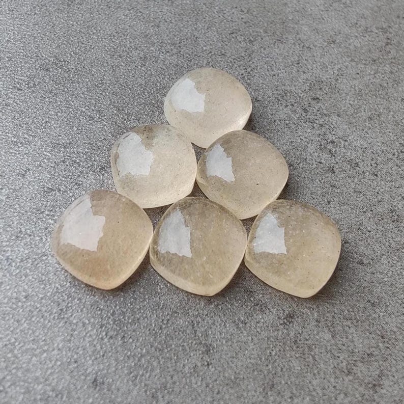 May include: Six light beige, square-shaped stones are arranged on a gray surface. Each stone has a rounded shape with a slightly reflective, white patch on one side. The stones appear smooth and translucent.