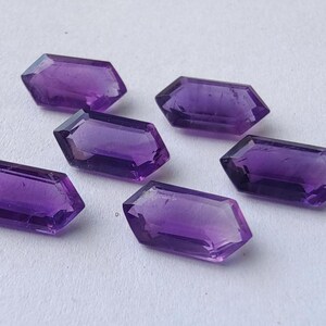 K&ouml;nnte beinhalten: Sechs facettierte Amethyst-Edelsteine in tiefem Lila liegen auf einer wei&szlig;en Oberfl&auml;che. Jeder Edelstein hat eine rechteckige Form mit abgewinkelten Seiten und eine polierte, reflektierende Oberfl&auml;che.