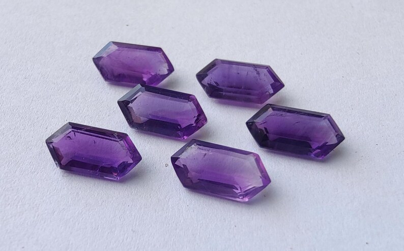 K&ouml;nnte beinhalten: Sechs facettierte Amethyst-Edelsteine in tiefem Lila liegen auf einer wei&szlig;en Oberfl&auml;che. Jeder Edelstein hat eine rechteckige Form mit abgewinkelten Seiten und eine polierte, reflektierende Oberfl&auml;che.