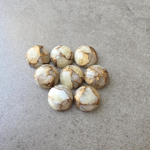 May include: Eight round, polished stones with a marbled appearance. The stones are primarily cream-colored with veins of gold and brown running through them. They are arranged on a gray surface.