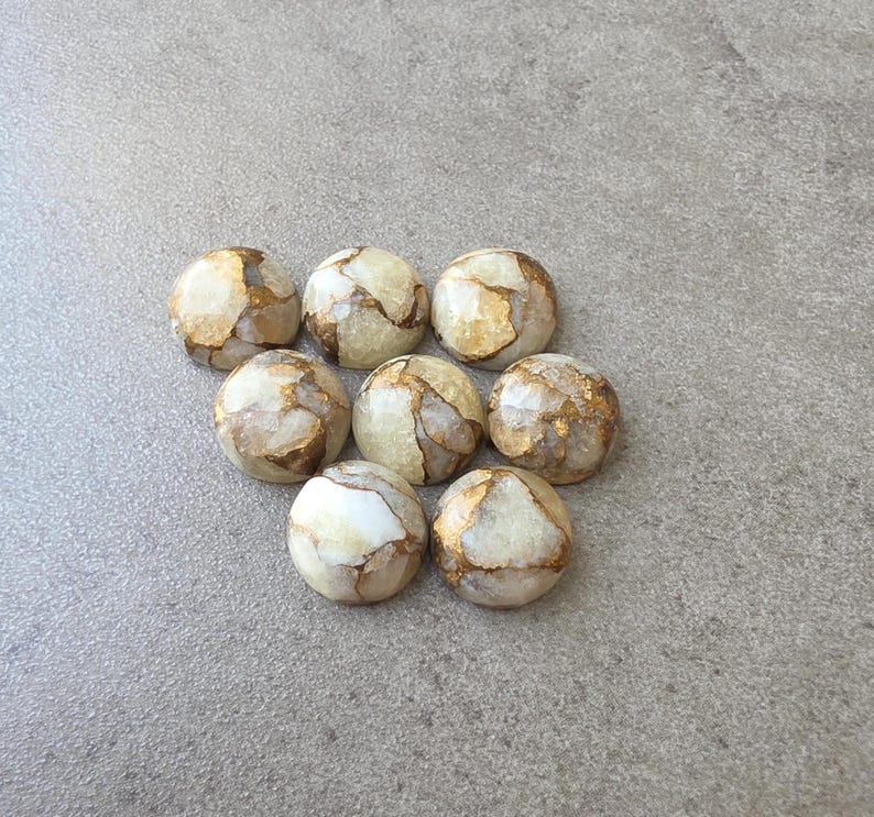 May include: Eight round, polished stones with a marbled appearance. The stones are primarily cream-colored with veins of gold and brown running through them. They are arranged on a gray surface.