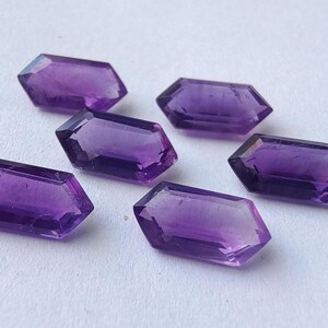 K&ouml;nnte beinhalten: Sechs facettierte Amethyst-Edelsteine in sechseckiger Form liegen auf einer wei&szlig;en Oberfl&auml;che. Die Edelsteine haben eine tiefviolette Farbe und sind mit flachen Ober- und Unterseiten versehen. Die Facetten fangen das Licht ein und erzeugen einen funkelnden Effekt. Jeder Edelstein ist etwa 1,5 cm lang.