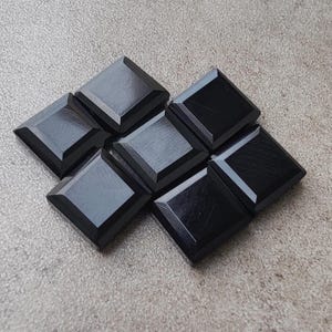 May include: A collection of eight square, black, beveled tiles arranged in a cluster. The tiles have a glossy finish and are set against a neutral, textured background. The arrangement suggests a mosaic or decorative element.