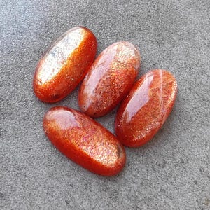 K&ouml;nnte beinhalten: Vier ovale Sonnenstein-Cabochons mit schimmerndem, irisierendem Finish. Die Steine sind eine Mischung aus Orange, Rot und Gold.