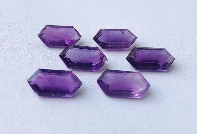 K&ouml;nnte beinhalten: Sechs facettierte, sechseckige Amethyst-Edelsteine. Die Edelsteine haben eine tiefviolette Farbe und sind auf einer wei&szlig;en Oberfl&auml;che angeordnet. Die Facetten fangen das Licht ein und erzeugen Glanzlichter und Schatten. Geeignet f&uuml;r die Schmuckherstellung.