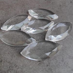 Könnte beinhalten: Sechs klare Glasprismenformen, die an Tropfen erinnern, liegen auf einer grauen Oberfläche. Die Prismen sind facettiert und reflektieren Licht.
