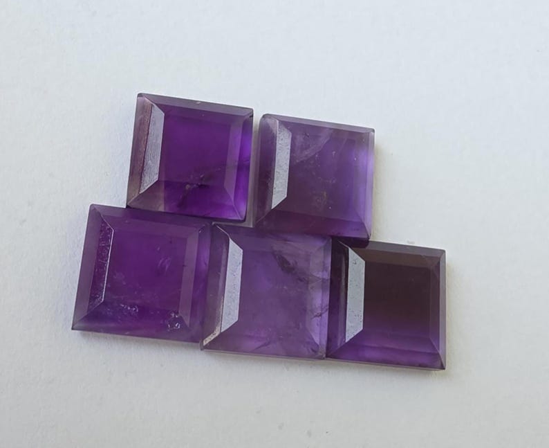 Peut inclure: Cinq pierres pr&eacute;cieuses d'am&eacute;thyste taill&eacute;es en carr&eacute;, de diff&eacute;rentes nuances de violet. Les pierres sont dispos&eacute;es en l&eacute;ger chevauchement, mettant en valeur leurs surfaces facett&eacute;es et leur couleur riche. Les pierres sont taill&eacute;es avec pr&eacute;cision.