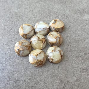 May include: A collection of eight round, polished stones. The stones have a creamy white base color with veins of gold and brown running throughout. The stones are arranged on a gray surface.