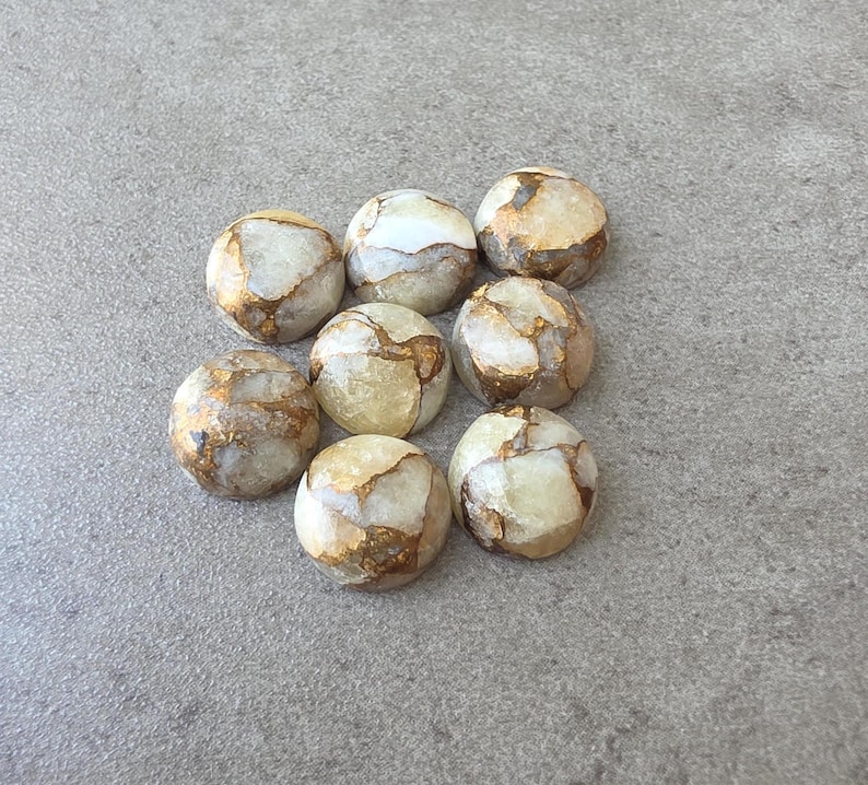 May include: A collection of eight round, polished stones. The stones have a creamy white base color with veins of gold and brown running throughout. The stones are arranged on a gray surface.