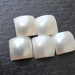 Pode incluir: Cinco cabochões quadrados, esbranquiçados, iridescentes. Cada um tem cantos arredondados e um brilho perolado, refletindo a luz com variações sutis de cor. Os cabochões estão dispostos em um padrão ligeiramente sobreposto sobre uma superfície escura texturizada.