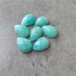 Op de afbeelding: Zeven gefacetteerde, druppelvormige edelstenen in tinten turquoise en aqua. De edelstenen zijn in een cirkelvormig patroon gerangschikt, elk met een gepolijst, reflecterend oppervlak. Op een neutrale grijze achtergrond.