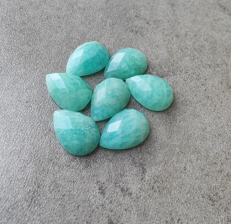 Op de afbeelding: Zeven gefacetteerde, druppelvormige edelstenen in tinten turquoise en aqua. De edelstenen zijn in een cirkelvormig patroon gerangschikt, elk met een gepolijst, reflecterend oppervlak. Op een neutrale grijze achtergrond.