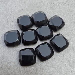 May include: Eight black, square-cut gemstones with faceted edges are displayed on a grey surface. The gemstones have a polished, reflective finish. The cushion cut and dark colour suggest they are suitable for jewellery design or crafting.