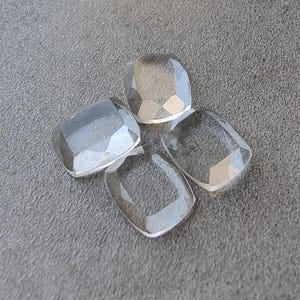 Może przedstawiać: Cztery przezroczyste, fasetowane, kwadratowe kamienie szlachetne ułożone na szarej powierzchni. Kamienie mają lekko zaokrąglony kształt i są przezroczyste. Fasetki tworzą efekt iskrzenia. Każdy kamień ma około 1,5 cm.