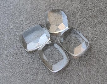 May include: Four clear, faceted, square-shaped gemstones are arranged on a gray surface. The gemstones have a slightly rounded shape and are transparent, allowing light to pass through. The facets create a sparkling effect.