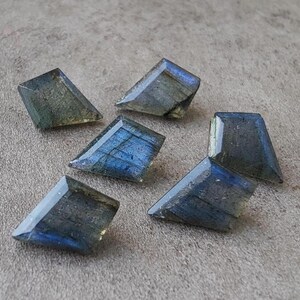 May include: Six trapezoid-shaped labradorite gemstones are displayed on a grey surface. The stones exhibit a mix of blue, green, and grey hues, with an iridescent shimmer. Each stone is faceted.